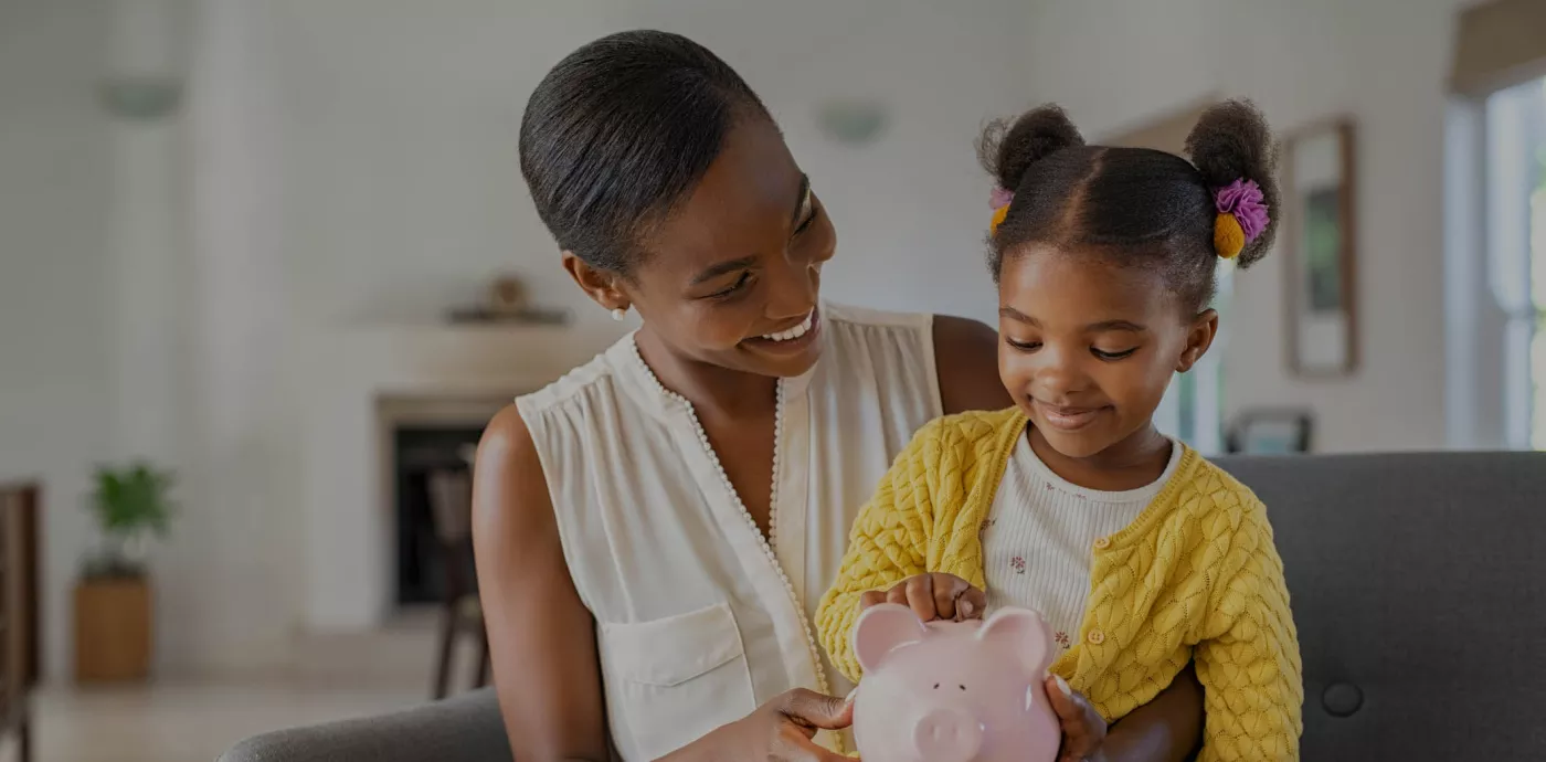 Mother holding daughter as she puts a coin in her piggy bank